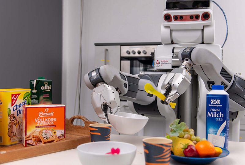 A robot holds a spoon and a bowl. In front of him is a table with cornflakes, milk, fruits and more.