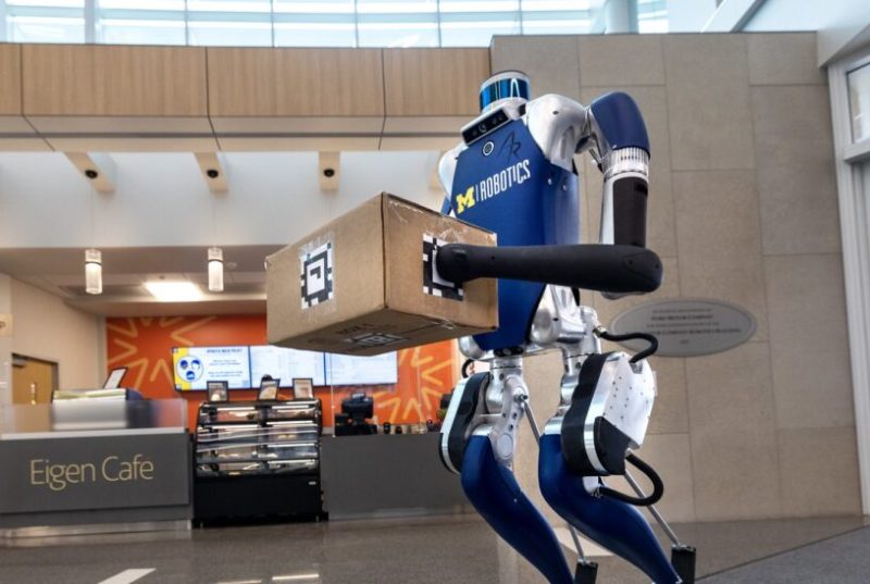 A humanoid robot is holding a cardboard box between both hands. The robot has the University of Michigan logo on its chest.